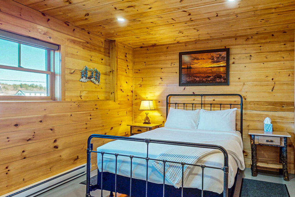 Cozy bedroom in Brakenhold with wooden walls, a metal bed frame, and a scenic wall art, featuring natural light from a window, enhancing the family-friendly atmosphere of King's Landing accommodations.